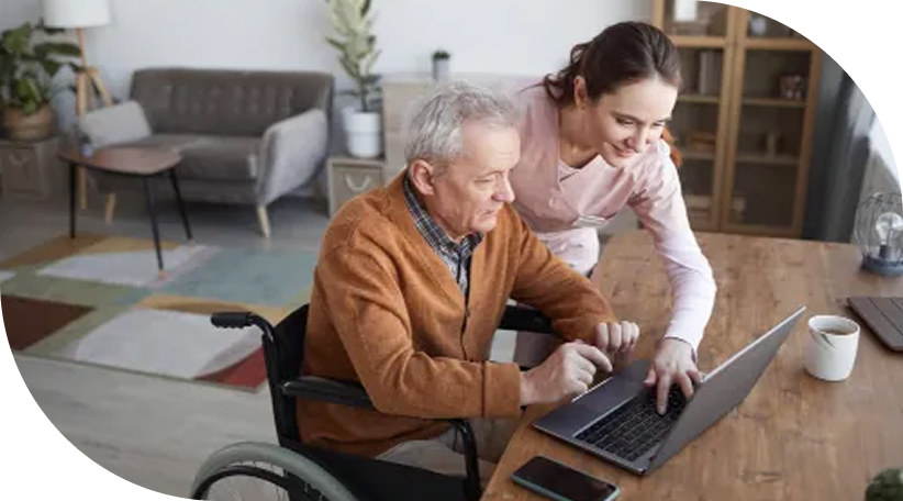 Portrait of senior man in wheelchair using laptop at retirement home with nurse assisting him