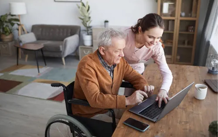 Portrait of senior man in wheelchair using laptop at retirement home with nurse assisting him